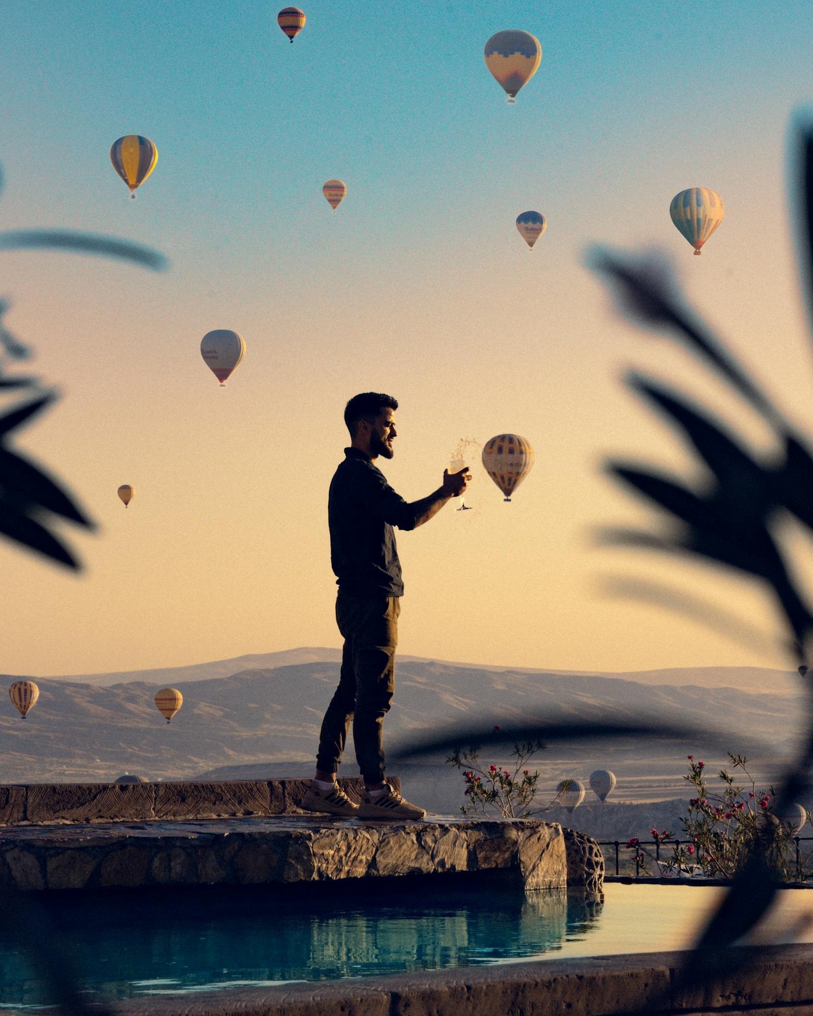 Man standing by pool holding wine glass, enjoying view of hot air balloons in the sky at sunset.